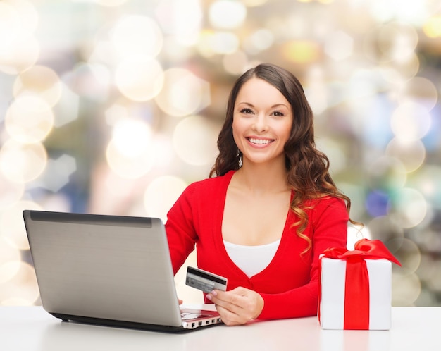 noël, vacances, technologie et concept de shopping - femme souriante avec carte de crédit, boîte-cadeau et ordinateur portable sur fond de lumières