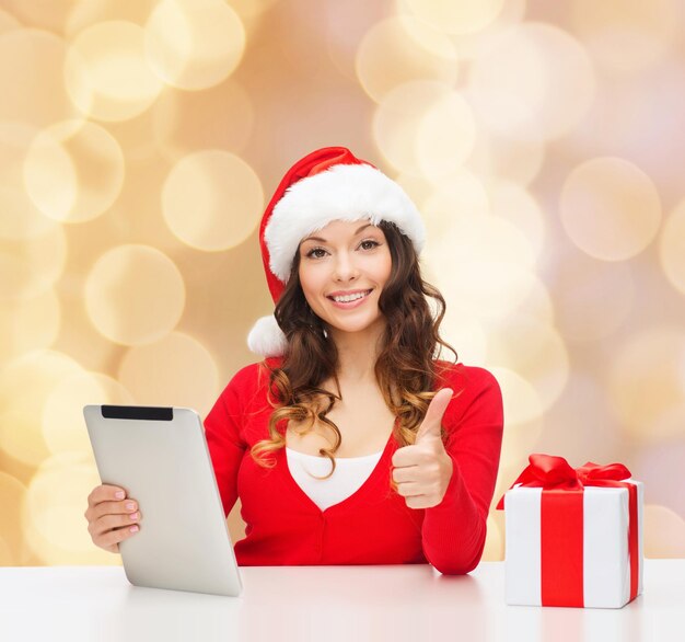 Noël, vacances, technologie, geste et concept de personnes - femme souriante en bonnet d'assistance avec boîte-cadeau et ordinateur tablette montrant les pouces vers le haut sur fond de lumières beiges