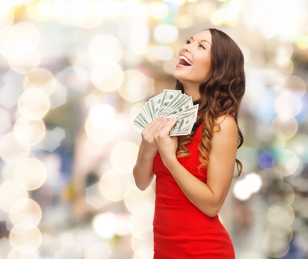 Noël, vacances, vente, banque et concept de personnes - femme souriante en robe rouge avec de l'argent en dollars américains sur fond de lumières