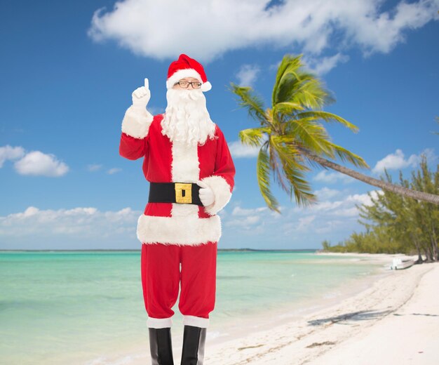 Photo noël, vacances, voyage, geste et concept de personnes - homme en costume de père noël pointant le doigt vers le haut sur fond de plage tropicale