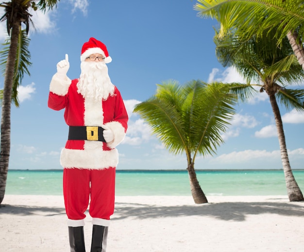 noël, vacances, voyage, geste et concept de personnes - homme en costume de père noël pointant le doigt vers le haut sur fond de plage tropicale