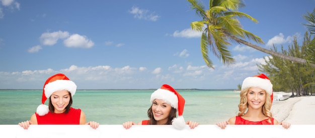 noël, x-mas, personnes, publicité et concept de vente - femmes heureuses en bonnet d'assistance avec tableau blanc vierge sur fond de plage tropicale