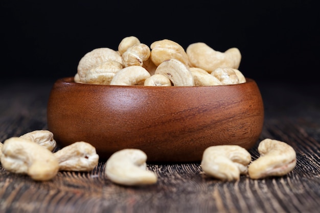 Noix de cajou sur une vieille table en bois et dans un bol en bois, gros plan d'un grand nombre de noix de cajou sur une table et sur une surface en bois dans une assiette en bois