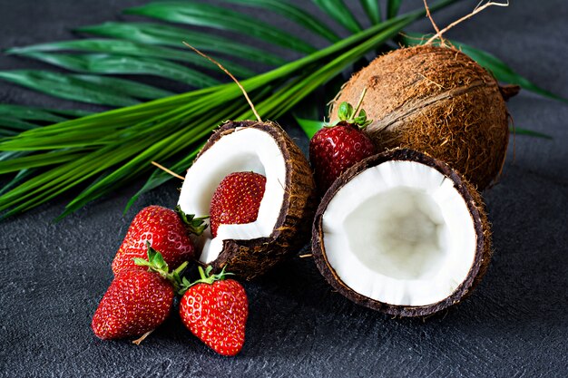 Noix de coco mûre avec des feuilles de palmier et des fraises fraîches mûres dans une demi-noix de coco sur un fond sombre. Fruits d'été, alimentation saine et délicieux dessert cru. Concept d'aliments sains, nourriture végétalienne.