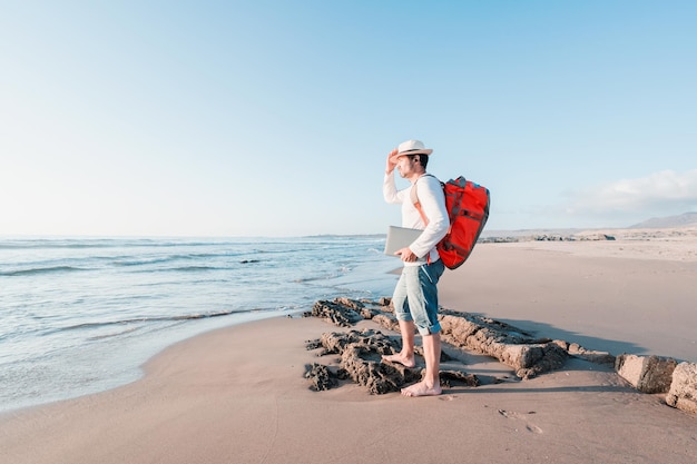 Nomade numérique avec un sac à dos et un ordinateur portable sur la plage regardant le coucher du soleil