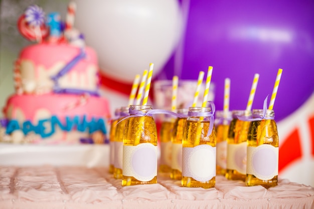 De nombreuses bouteilles de jus de pomme, des étiquettes spéciales, des pailles blanches et jaunes, un gros gâteau rose et des ballons blancs et violets