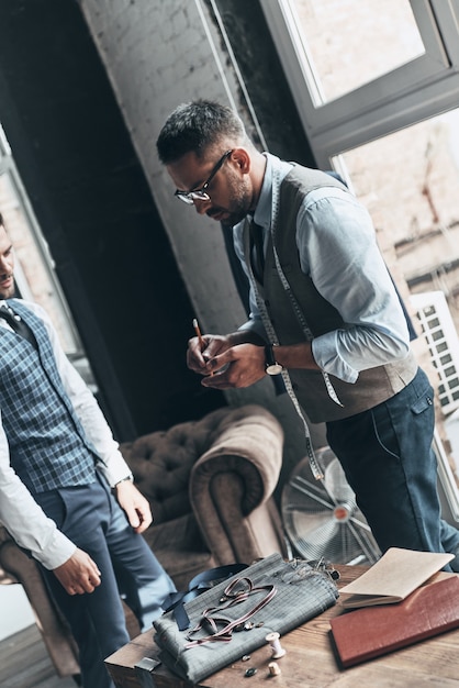 En notant attentivement. Deux jeunes hommes à la mode ayant une discussion en se tenant debout en atelier