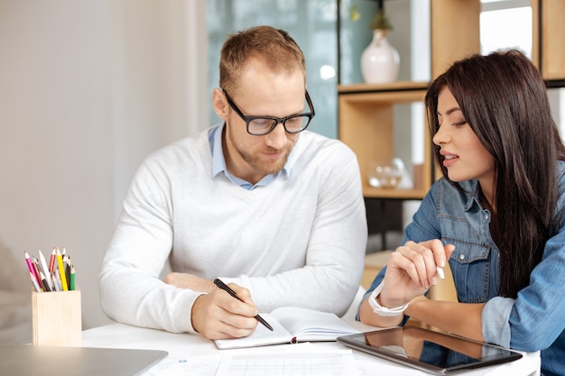 Noter un plan. Intelligent bel homme gentil assis avec son collègue et regardant le cahier tout en élaborant un plan pour les travaux futurs