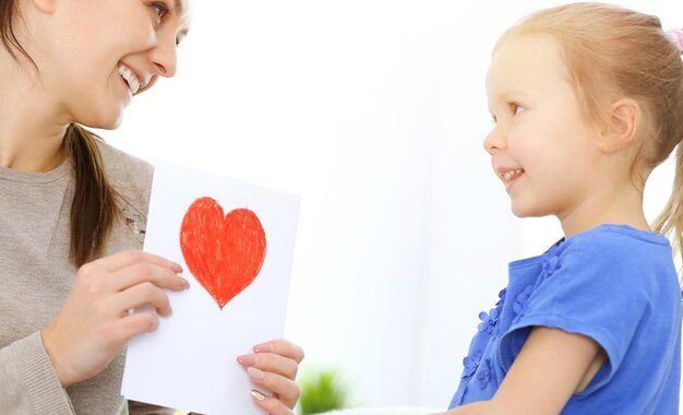 Notion de fête des mères. La petite fille félicite maman et lui donne une carte postale en forme de coeur rouge. Maman et fille heureuse souriante et étreignant. Plaisir en famille et vacances.