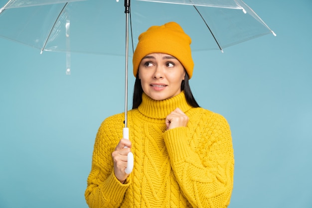 Notion de jour de pluie. Portrait d'une belle femme triste tenant un parapluie transparent et levant isolé sur un mur bleu. Stock photo