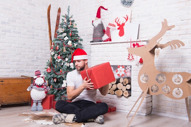 Notion de lendemain de Noël. Macho tient une boîte présente à l'arbre de Noël. Préparation des cadeaux de Noël et du nouvel an. Joyeuses fêtes de fin d'année. Man in santa hat s'asseoir sur le sol à la cheminée.