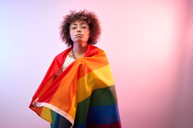 Notion LGBT. Fille caucasienne positive aux cheveux bouclés afro tenant le drapeau arc-en-ciel isolé en studio