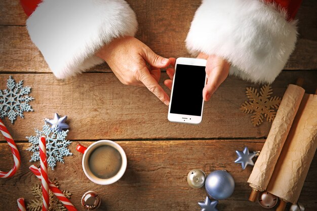 Notion de Noël. Le père Noël prend le téléphone intelligent dans les mains sur une table en bois en gros plan