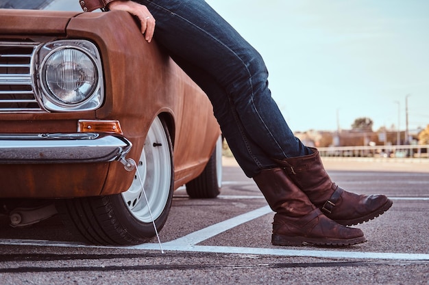 Notion de style campagnard. Photo recadrée d'un homme en jeans et bottes s'appuyant sur une voiture rétro dans le parking de la ville.