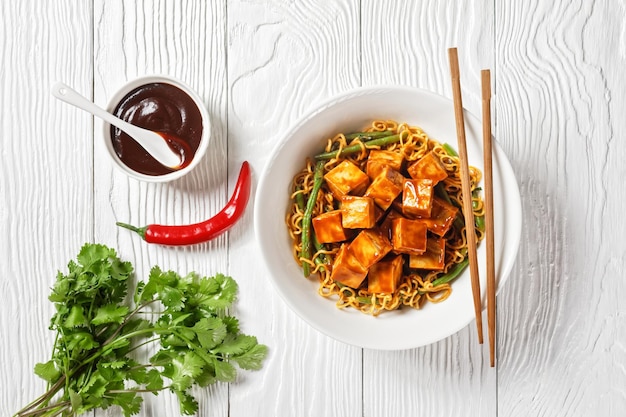 Nouilles asiatiques sucrées et épicées avec du tofu croustillant au four hoisin et des haricots verts dans un bol blanc avec des baguettes vue horizontale d'en haut à plat