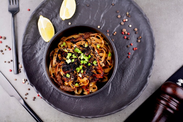 Nouilles au wok aux crevettes, dans une assiette noire décorée de petits pois, sur fond gris. Le concept de la cuisine asiatique.