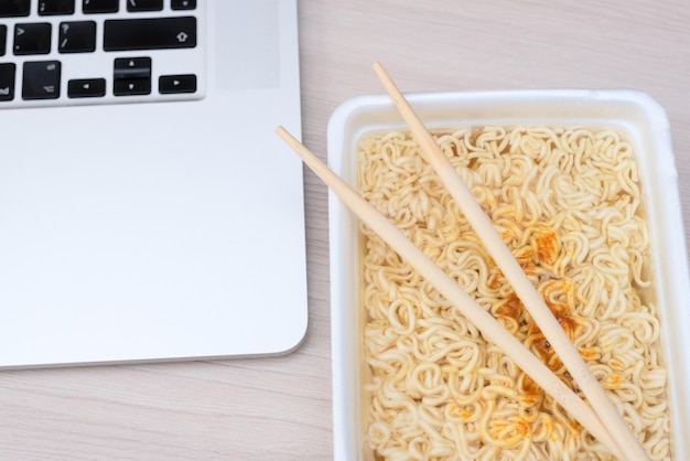 Nouilles frisées chinoises instantanées dans des emballages à emporter avec des bâtons de bois et un ordinateur portable sur la table de bureau Mise au point sélective Nourriture asiatique et concept de livraison Alimentation malsaine