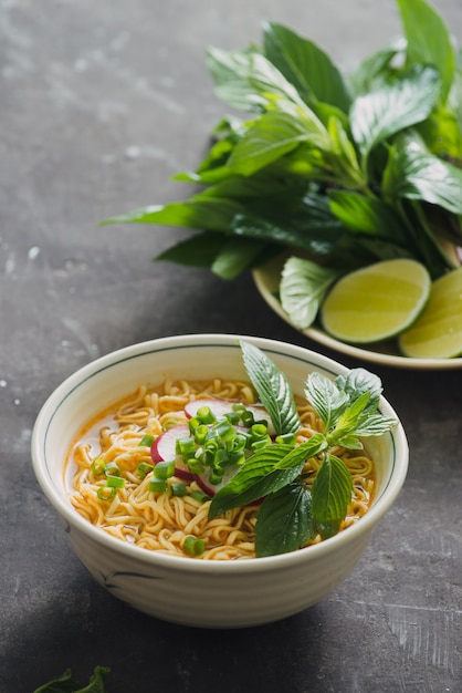 Nouilles instantanées dans un bol avec des herbes fraîches, garniture de coriandre et basilic asiatique, citron, citron vert sur fond de pierre sombre