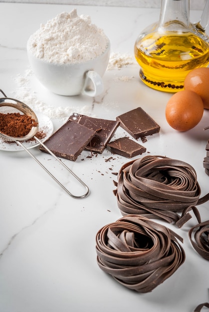 Nouilles de pâtes au chocolat crues non préparées, avec des ingrédients pour la cuisine - chocolat, cacao, farine, œufs, huile. Sur une table en marbre de cuisine blanche. fond