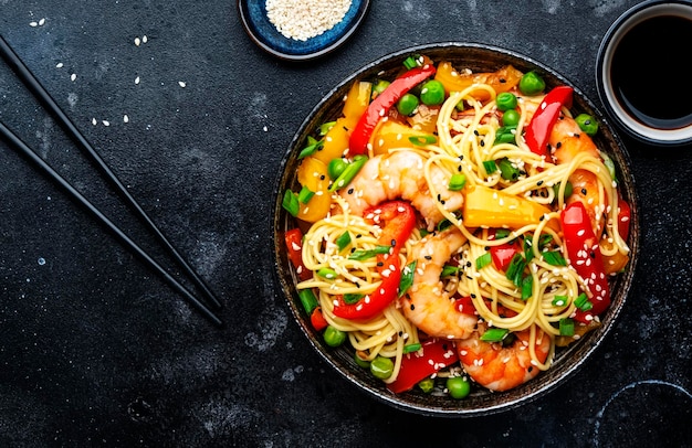 Nouilles sautées aux crevettes paprika rouge et jaune pois verts ciboulette et graines de sésame bol plat de cuisine asiatique cuisine en pierre noire fond de table vue de dessus