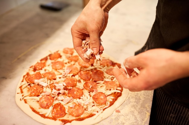 nourriture, cuisine, cuisine italienne, personnes et concept de cuisine - mains de cuisinier ajoutant du jambon à la pizza au salami à la pizzeria