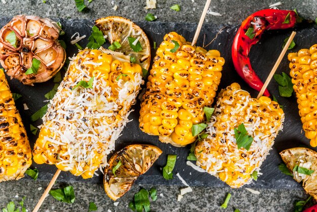 Photo la nourriture d'été. idées de soirées barbecue et grillades. maïs grillé grillé au feu. avec une pincée de fromage (élotes mexicaines), du piment fort et du citron. sur une table en pierre sombre. vue rapprochée du haut
