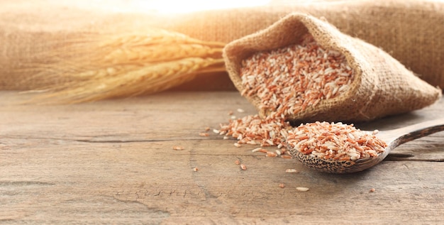 Photo nourriture saine gros plan cuillère de grains de riz entiers biologiques rouges et sac brun sur table en bois avec oreille de paddy sur fond brillant