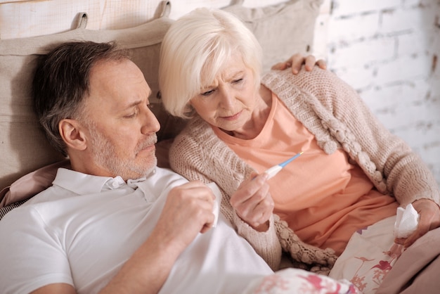 Nous avons de la fièvre. Vue de dessus du couple de personnes âgées allongé sur le lit et vérifier l'heure sur le thermomètre.