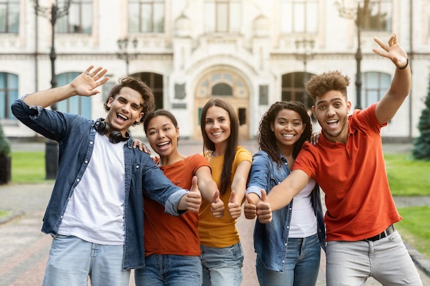 Nous recommandons un groupe d'étudiants multiethniques joyeux gesticulant les pouces vers la caméra