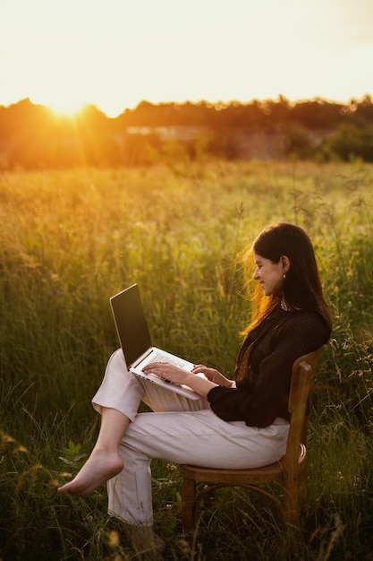 Nouveau concept de bureau Fille élégante à la mode travaillant sur un ordinateur portable et assise sur une chaise rustique dans un champ d'été ensoleillé au coucher du soleil Travail à distance avec des protocoles de distanciation sociale et de sécurité