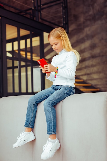 Nouveau gadget. Fille blonde concentrée assise sur un canapé et discutant en ligne avec son amie