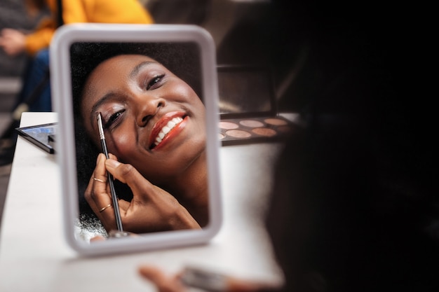 Nouveau maquillage. Femme afro-américaine souriante portant un haut orange mettant des ombres à paupières sur ses paupières