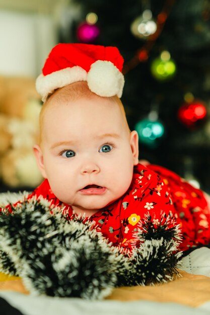 Un nouveau-né avec un chapeau de Père Noël rouge est allongé sur le lit près de l'arbre de Noël.