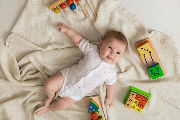 Un nouveau-né mignon dans des vêtements légers joue avec des jouets éducatifs allongés sur le sol sur une couverture en lin gris. vue de dessus. produits pour enfants. enfance et maternité heureuses. espace pour le texte