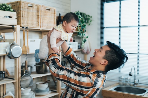 un nouveau père asiatique heureux soulève son bébé innocent tout en jouant avec lui dans une cuisine moderne et lumineuse à la maison.