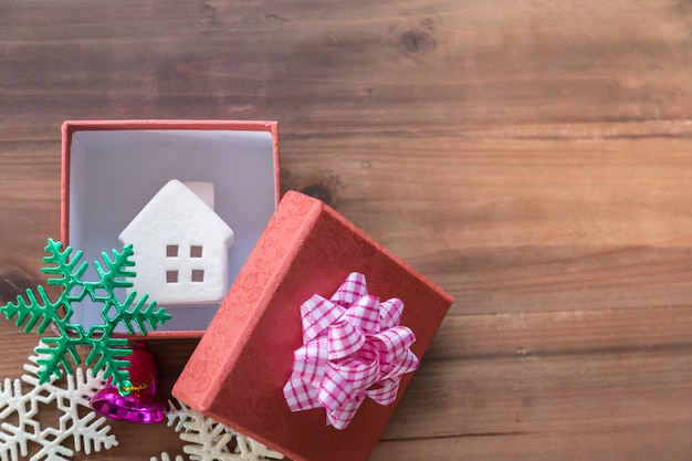 Nouvel an, famille, Noël et concept d'assaisonnement de vacances. Gros plan de l'icône de la maison dans une boîte cadeau rouge avec un beau ruban avec une cloche violette et des ornements de neige colorés sur planche de bois avec espace de copie.