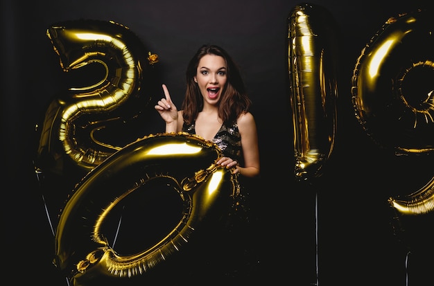 Nouvelle année. Femme avec des ballons célébrant à la fête. Portrait de belle fille souriante en robe brillante jetant des confettis, s'amusant avec des ballons d'or 2019 sur fond. Haute résolution.