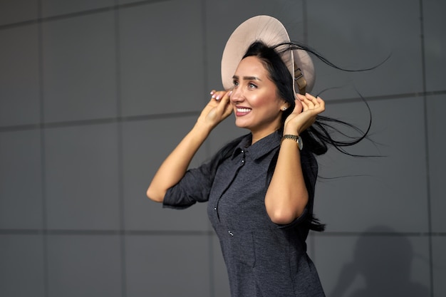 Nouvelle collection jeunesse de vêtements pour femmes. Portrait agrandi en plein air jolie fille drôle aux cheveux longs portant une robe oversize grise des jeunes et un chapeau élégant en journée venteuse.