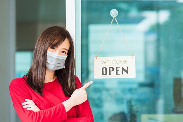 Une nouvelle jeune femme asiatique normale porte un masque facial protège le doigt pointé heureux pour remarquer l'étiquette de panneau de bois "WELCOME OPEN" accrochée à travers la porte vitrée de la boutique, l'entreprise s'ouvre après la pandémie de coronavirus