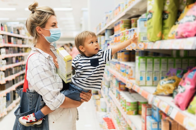 Nouvelle norme dans les achats. Jeune mère portant un masque de prévention et son petit fils dans un supermarché pendant une pandémie de virus.