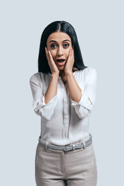 Nouvelles étonnantes. Jeune femme surprise en chemise blanche tenant les mains sur son visage et regardant la caméra en se tenant debout sur fond gris