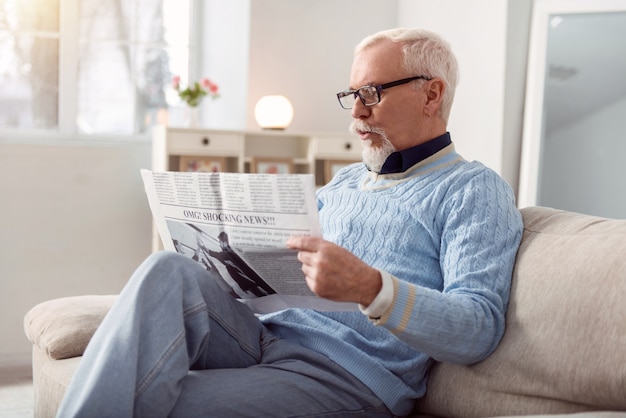 Nouvelles fraîches. Bel homme barbu assis confortablement sur le canapé dans le salon et lisant un journal, surpris par les nouvelles qu'il contient