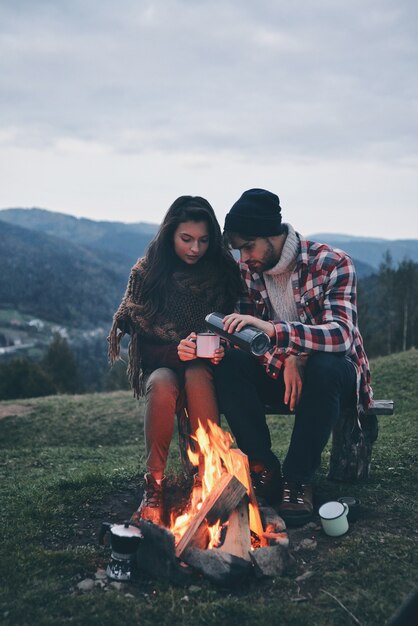 Nuit romantique. Beau jeune couple dégustant des boissons chaudes tout en se réchauffant près du feu de camp dans les montagnes