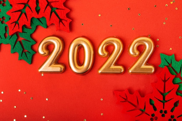 Numéros d'or rose 2022 sur fond rouge. Belles feuilles de houx décoratives autour. Fond de nouvel an.