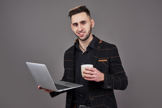 Occupé homme barbu en vêtements d'affaires parler par smartphone et à l'aide d'un ordinateur portable tout en tenant une tasse de café à la main sur gris