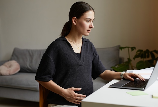 Occupé jeune femme caucasienne enceinte assise à table dans le salon et utilisant un ordinateur portable tout en travaillant à distance