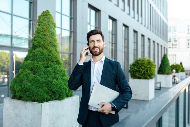 Occupé souriant confiant jeune homme d'affaires caucasien avec barbe en costume avec appel d'ordinateur portable par smartphone