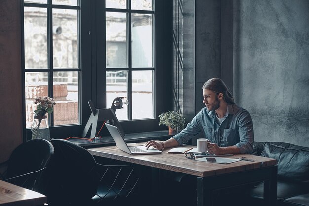 Occupé à travailler au bureau. Jeune homme réfléchi travaillant à l'aide d'un ordinateur et tenant une tasse