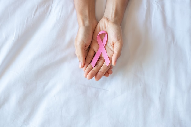 Octobre Mois De Sensibilisation Au Cancer Du Sein, Main De Femme Adulte Tenant Un Ruban Rose Sur Fond Rose Pour Soutenir Les Personnes Vivant Et Malades. Concept De La Journée Internationale Des Femmes, Des Mères Et Du Monde Contre Le Cancer