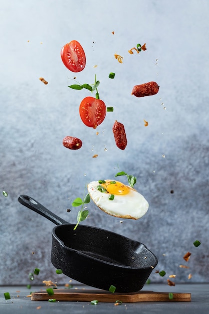 Oeuf au plat volant en mouvement. Oeuf au plat et saucisses fumées sur une poêle en fonte avec sel et assaisonnement, oignons verts et microgreen sur fond moderne en béton gris